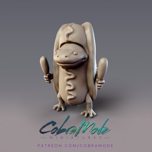 May include: A tan-colored figurine of a frog wearing a hot dog bun costume and holding two drumsticks. The figurine has a whimsical design with a smiling face and detailed features. The text "CobraMode Miniatures" and a Patreon link are visible.