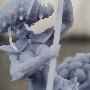 May include: A gray 3D printed miniature of a female figure with a spear, wearing armor and a helmet with spikes. The figure is standing on a base of smaller, rounded shapes.