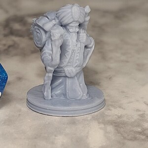 May include: A gray 3D printed miniature of a figure wearing a turban and carrying a staff and a backpack. The figure is standing on a round base. A blue 20-sided die is in the foreground.