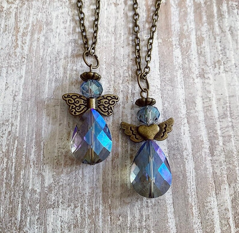 May include: Two angel pendant necklaces. Each necklace features a teardrop-shaped, iridescent crystal pendant with bronze-colored wings and a chain. The crystals reflect blue and purple hues. The necklaces hang against a light-colored, textured background.