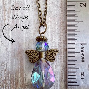 May include: A pendant necklace featuring a crystal angel with scroll wings. The angel pendant has a large, iridescent teardrop-shaped crystal body, with a smaller crystal head and ornate bronze-colored wings. The chain is bronze-colored. Text on the image reads "Scroll Wings Angel".