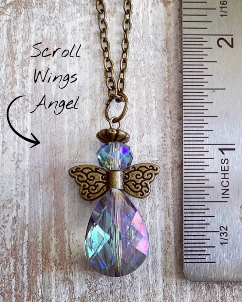 May include: A pendant necklace featuring a crystal angel with scroll wings. The angel pendant has a large, iridescent teardrop-shaped crystal body, with a smaller crystal head and ornate bronze-colored wings. The chain is bronze-colored. Text on the image reads "Scroll Wings Angel".
