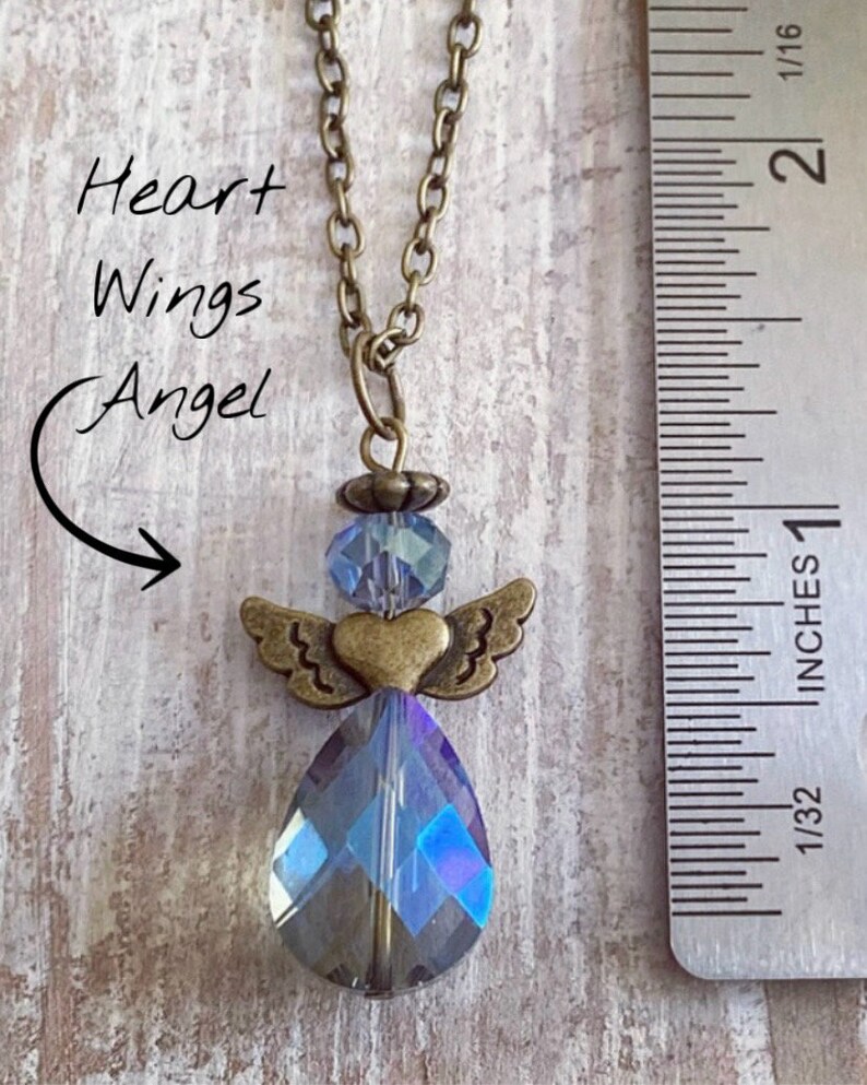 May include: A bronze-toned necklace featuring a pendant in the shape of an angel. The angel has a heart-shaped body, small wings, and a large, iridescent teardrop-shaped crystal. The text "Heart Wings Angel" is written on the left side of the image.