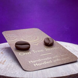 May include: A pair of dark brown coffee bean stud earrings. The earrings are displayed on a brown card with the text "Owl Tree Jewelry" and "Handmade with love, Handled with care". The background is a gradient of purple.