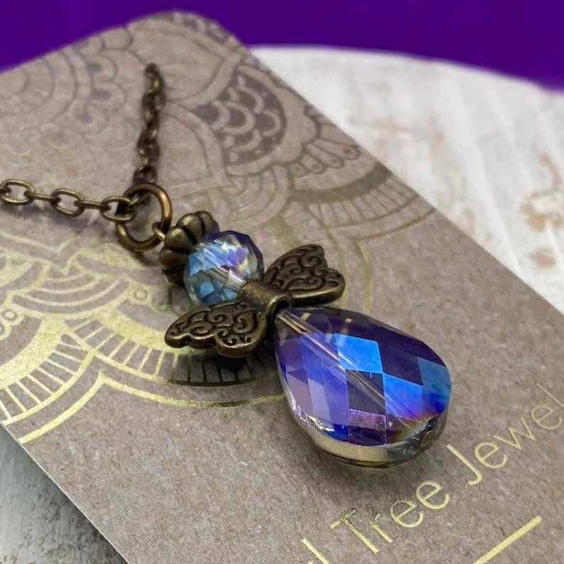 May include: A bronze-toned angel pendant necklace. The pendant features a faceted, iridescent blue teardrop crystal body, a smaller blue crystal head, and ornate bronze wings. The necklace chain is also bronze.