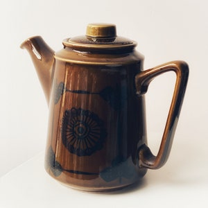 以下が含まれることがあります： 花柄の茶色の陶器製のティーポット。ティーポットには蓋と取っ手があります。デザインは黒と茶色です。