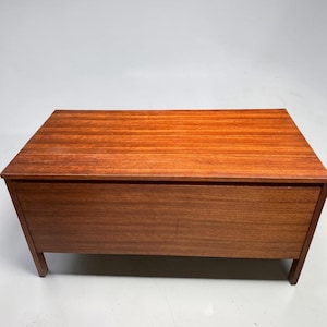 Könnte beinhalten: Eine rechteckige Holzkiste mit rotbraunem Finish. Die Truhe hat eine flache Oberseite und eine Frontplatte mit horizontalem Maserungsmuster. Sie wird von vier kurzen, rechteckigen Beinen getragen. Die Truhe scheint leer zu sein.