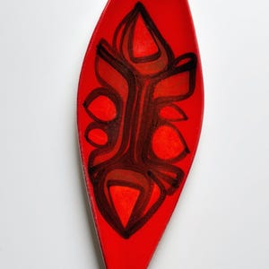 May include: A bright red, elongated ceramic dish with a glossy finish. The dish has a central abstract design in shades of brown and black. The design is made up of organic shapes and lines, creating a striking contrast against the red background.