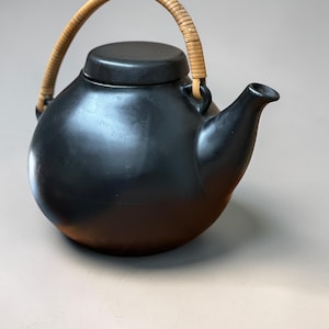 May include: A black ceramic teapot with a rounded body, a short spout, and a lid. The handle is made of woven light brown rattan. The teapot is designed for brewing tea and has a minimalist aesthetic.