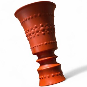Puede incluir: Un jarrón de cerámica naranja con una superficie texturizada. El jarrón tiene una parte superior acampanada y una base en espiral texturizada. El diseño incluye filas de pequeños puntos en relieve. El jarrón mide unos 25 cm de alto.