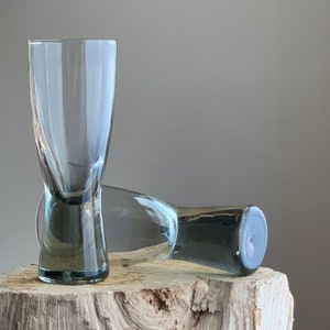 May include: Two clear glass shot glasses with a grey tint. The glasses are sitting on a piece of weathered wood.