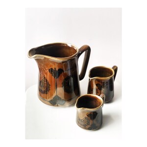 Peut inclure: Un ensemble de trois pichets en céramique brune avec un motif floral noir. Le plus grand pichet a une large ouverture et une poignée. Les deux plus petits pichets ont une ouverture plus petite et une poignée.