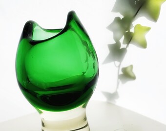 May include: A green glass vase with a unique, organic shape and a clear glass base. The vase is sitting on a white surface.