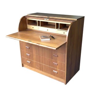 Könnte beinhalten: Ein Holzschreibtisch mit Rollladen und warmer brauner Oberfläche. Der Schreibtisch verfügt über eine ausziehbare Schreibfläche, drei Schubladen und mehrere kleine Fächer. Ein Notizbuch und ein Stift liegen auf der Schreibfläche. Der Schreibtisch ist ein klassisches Möbelstück.