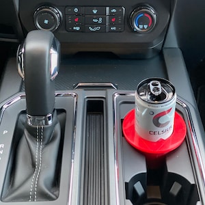 May include: A black car interior with a gear shift lever, a cup holder, and a can of Celsius energy drink in a red cup holder.