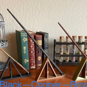 May include: Three wooden wand stands in black, copper, and bronze finishes. Each stand features a triangular base and a slot to hold a wand. The stands are displayed with wands on a wooden surface.