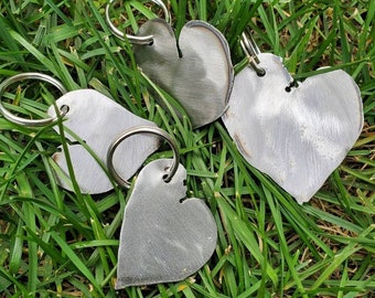 May include: A collection of heart-shaped metal keychains with silver key rings, resting on a bed of green grass. The keychains have a brushed metal finish, and each heart has a unique shape and texture.