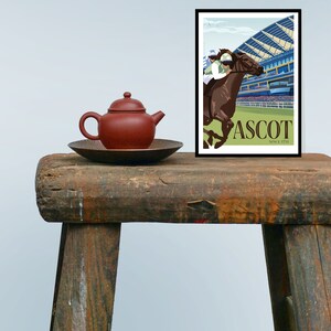 May include: A framed poster with a horse racing scene at Ascot. The artwork features a brown horse with a jockey, set against a backdrop of a grandstand. A small, red teapot and a dark brown plate sit on a rustic wooden stool.