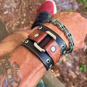 May include: A brown leather bracelet with a black leather strap and silver buckle. The bracelet has a distressed look and is worn on a person's wrist.