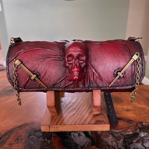 May include: A dark red leather clutch with a skull design and brass accents. The clutch has a chain strap and a buckle closure.