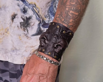 Puede incluir: Un brazalete de cuero marrón con un diseño decorativo en relieve y tachuelas de latón. El brazalete se lleva en la muñeca.