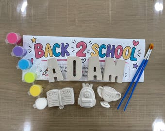 May include: A DIY painting kit featuring a white paper sign with the text BACK 2 SCHOOL, four plaster figures shaped like the letters ADAM, a book, a backpack, and a trophy. Includes a strip of six colorful paint pots and two blue paintbrushes.