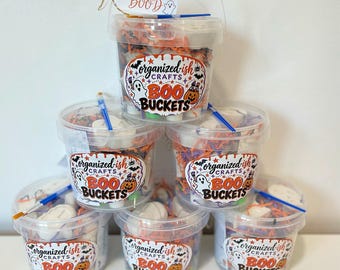 May include: Six clear plastic buckets filled with orange and purple paper shred, small white ceramic figures, and a blue-handled paintbrush. Each bucket features a label reading Organized-ish Crafts BOO BUCKETS with a ghost and pumpkin graphic.