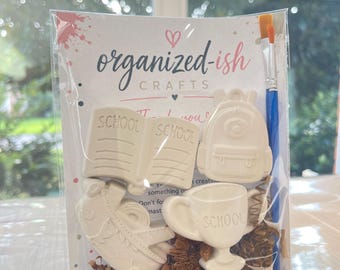 May include: A craft kit in a clear plastic bag featuring three white ceramic figures: an open book, a backpack, and a trophy, each embossed with the word SCHOOL. The kit includes a blue paintbrush and three small paint pots in blue, purple, and pink.
