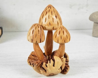 Può includere: Una scultura in legno intagliata a mano raffigurante tre funghi di diverse altezze. Ogni cappello di fungo presenta un motivo testurizzato a forma di diamante. I gambi sono lisci e marrone chiaro, su una base di radici di legno intagliate.