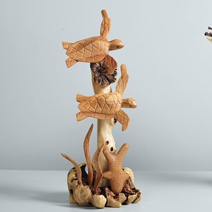 May include: A light brown wooden sculpture featuring two carved sea turtles attached to a central vertical branch. The base is composed of textured wood elements resembling coral and seaweed, with a small starfish-like shape at the bottom.