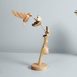 Puede incluir: Una escultura de madera de un colibrí posado en una rama, con una flor tallada en el extremo de la rama. La escultura está hecha de madera natural de color marrón claro.