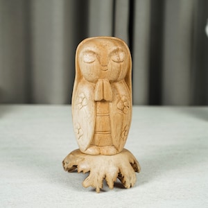 Könnte beinhalten: Eine hellbraune Holzfigur mit geschlossenen Augen und gefalteten Händen in Gebetshaltung. Die Figur weist geschnitzte Blumendetails an den Seiten auf und ist auf einem natürlichen, unregelmäßigen Holzsockel mit sichtbaren Wurzeln montiert.