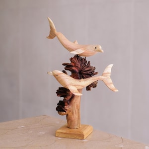 Könnte beinhalten: Eine Holzskulptur mit zwei geschnitzten Delfinen, die an einem zentralen Stück dunklem, strukturiertem Treibholz befestigt sind. Die Delfine sind hell gefärbt mit sichtbarer Holzmaserung und auf einem quadratischen Holzsockel montiert.
