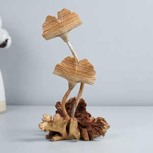 Könnte beinhalten: Eine dekorative Skulptur mit zwei geschnitzten Ginkgoblättern aus Holz an schlanken Stielen, die aus einem strukturierten, organischen Holzsockel ragen. Die Blätter zeigen sichtbare Holzmaserungen in hellbraunen Tönen, der Sockel ist dunkelbraun.