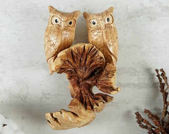 May include: Two carved wooden owls perched on a tree stump. The owls have black eyes and detailed feathers. The tree stump is brown and has a rough texture.