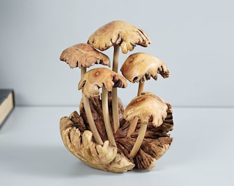 Può includere: Una scultura decorativa raffigurante cinque funghi in legno intagliato con cappelli marrone chiaro testurizzati e gambi lisci. I funghi sono disposti a grappolo, emergendo da una base costituita da un pezzo di legno naturale marrone scuro.