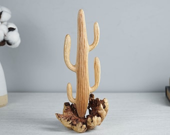 May include: A light brown wooden sculpture shaped like a saguaro cactus with a tall central column and four curved arms. The cactus is mounted on a textured, dark brown natural wood burl base that shows organic knots and irregular grain patterns.
