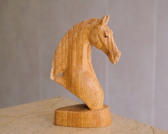 May include: A light brown wooden sculpture of a horse head and neck, featuring visible wood grain patterns and a textured mane. The sculpture is mounted on a solid, oval-shaped wooden base, capturing the profile of the animal in a static pose.