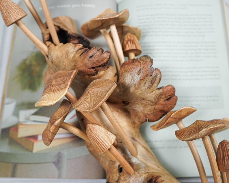 May include: A collection of carved wooden mushrooms with detailed caps and smooth stems, attached to a piece of natural, textured driftwood. The mushrooms vary in size and shape, featuring light brown wood grain patterns throughout the sculpture.