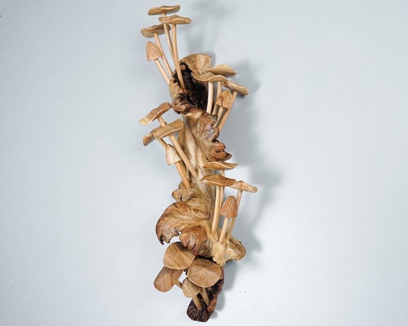 May include: A vertical wooden sculpture featuring a cluster of carved light brown mushrooms emerging from a textured, gnarled piece of natural wood. The mushrooms vary in size and shape, with some having flat caps and others having pointed, conical tops.