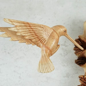 May include: A wooden hummingbird sculpture with detailed wings and a long beak, perched on a branch with carved leaves.
