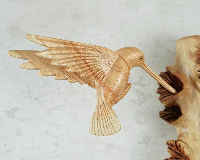 May include: A wooden hummingbird sculpture with detailed wings and a long beak, perched on a branch with carved leaves.