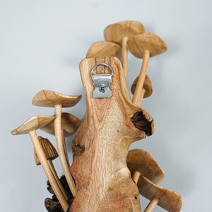 May include: A wall-mounted wooden sculpture featuring multiple carved mushroom shapes attached to a central piece of textured, light brown wood. A silver-colored metal D-ring hanger is securely fastened to the top center of the wooden base.