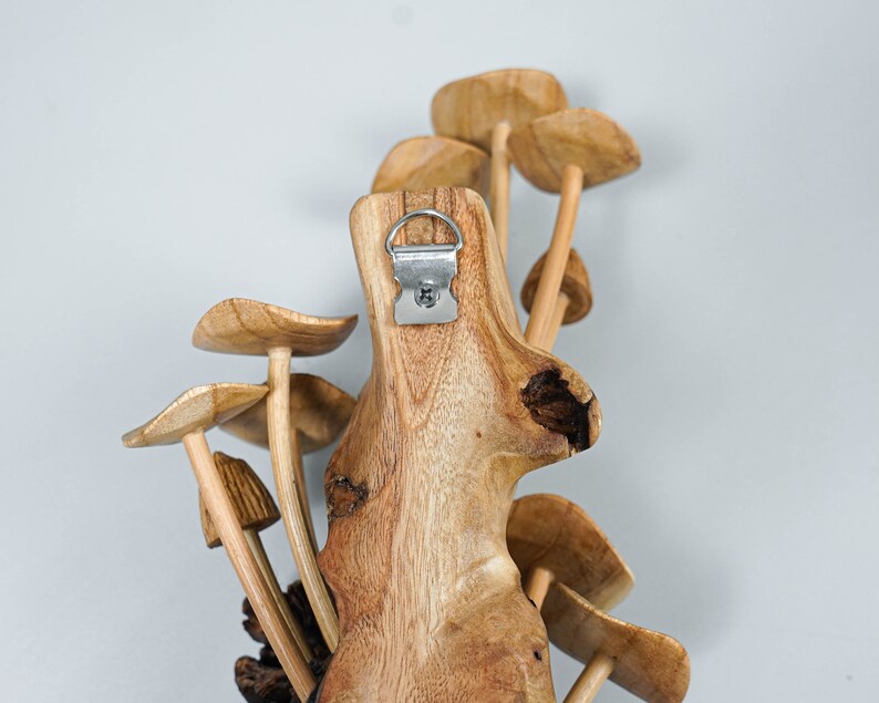 May include: A wall-mounted wooden sculpture featuring multiple carved mushroom shapes attached to a central piece of textured, light brown wood. A silver-colored metal D-ring hanger is securely fastened to the top center of the wooden base.