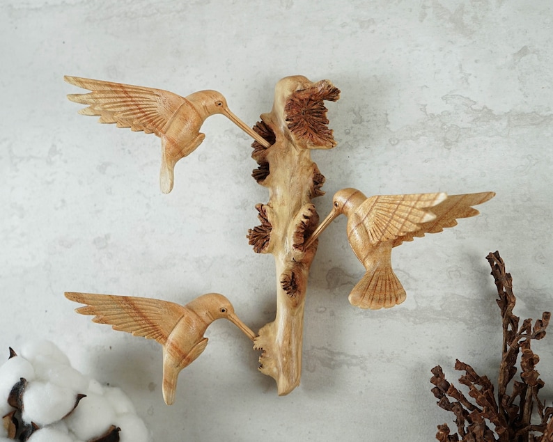 May include: Three wooden hummingbirds carved in detail, perched on a branch with a natural wood finish. The birds are facing different directions, with their wings spread out.