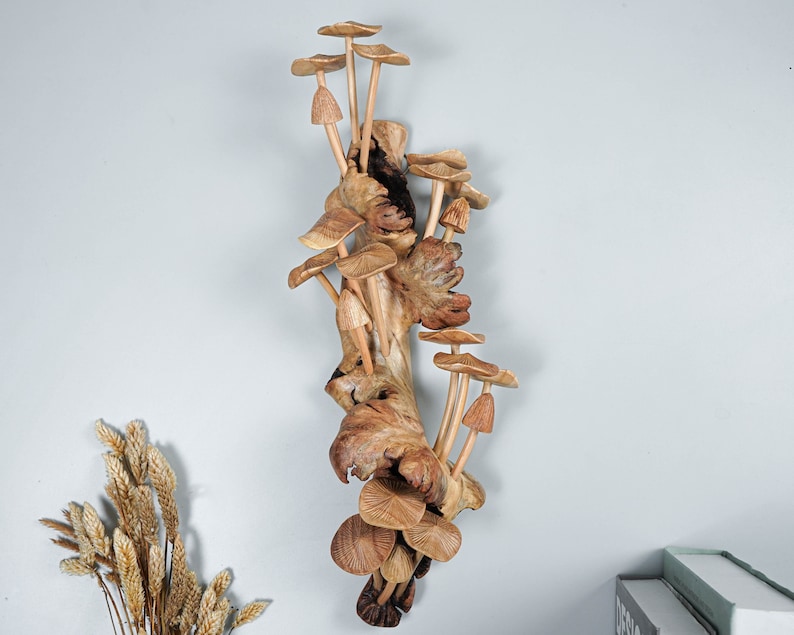 May include: A vertical wall sculpture featuring a cluster of carved wooden mushrooms attached to a piece of textured, gnarled driftwood. The mushrooms vary in size and shape, displaying natural wood grain patterns in shades of light and dark brown.