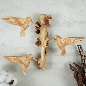 May include: Wooden wall art featuring three carved hummingbirds perched on a branch. The birds are painted in natural wood tones and have intricate details.