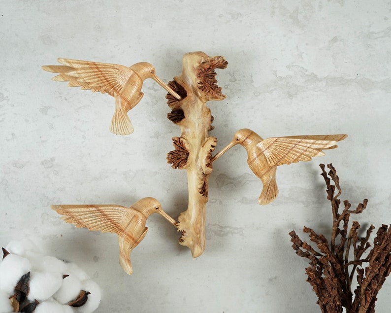 May include: Wooden wall art featuring three carved hummingbirds perched on a branch. The birds are painted in natural wood tones and have intricate details.