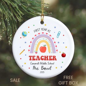 Peut inclure: Ornement de No&euml;l rond en c&eacute;ramique blanche avec un arc-en-ciel, une pomme et des graphiques scolaires. Le texte comprend "First Year as Teacher", le nom de l'&eacute;cole, le nom de l'enseignant et l'ann&eacute;e 2025. Une ficelle dor&eacute;e est attach&eacute;e.