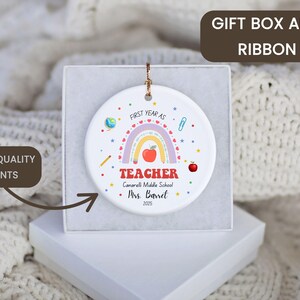 Peut inclure: Ornement en c&eacute;ramique blanc avec un arc-en-ciel, une pomme et le texte "FIRST YEAR AS TEACHER". L'ornement est pr&eacute;sent&eacute; dans un coffret cadeau blanc. Le texte comprend &eacute;galement "Canarelli Middle School" et "Mrs. Barret 2025."